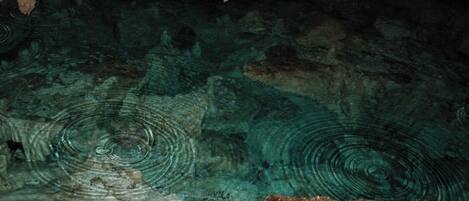 A cave interior with stalactites and stalagmites, clear water, and a large rock in the foreground.