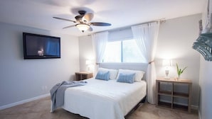 A bedroom with a bed, a ceiling fan, a TV mounted on the wall, a window with curtains, bedside tables with lamps, and a small plant on one table. - Stay a While - 3 min walk to private beach access!

 (Destin)
