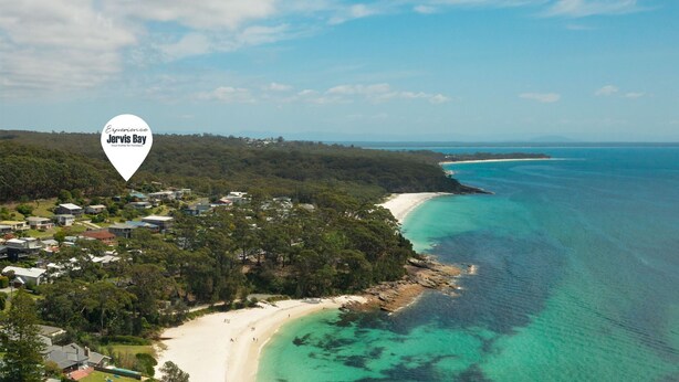 An aerial view of Jervis Bay with a coastal beach, residential area, and clear turquoise waters.