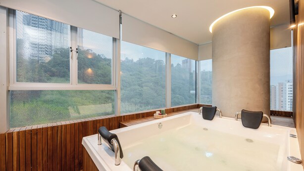 A modern bathroom with a large bathtub, wooden paneling, and large windows offering views of greenery and buildings. - 3BR Penthouse with Jacuzzi & A/C in El Poblado (Medellín)