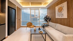 A modern living room with a sofa, a coffee table, and a chair. There is a large window with a city view, a wooden wall with a framed artwork, and a kitchen area with a refrigerator. - 3BR Penthouse with Jacuzzi & A/C in El Poblado (Medellín)