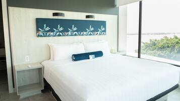 Una habitación de hotel moderna con una cama grande, una mesita de noche, un cuadro colgado en la pared y vistas a zonas verdes a través de amplias ventanas.