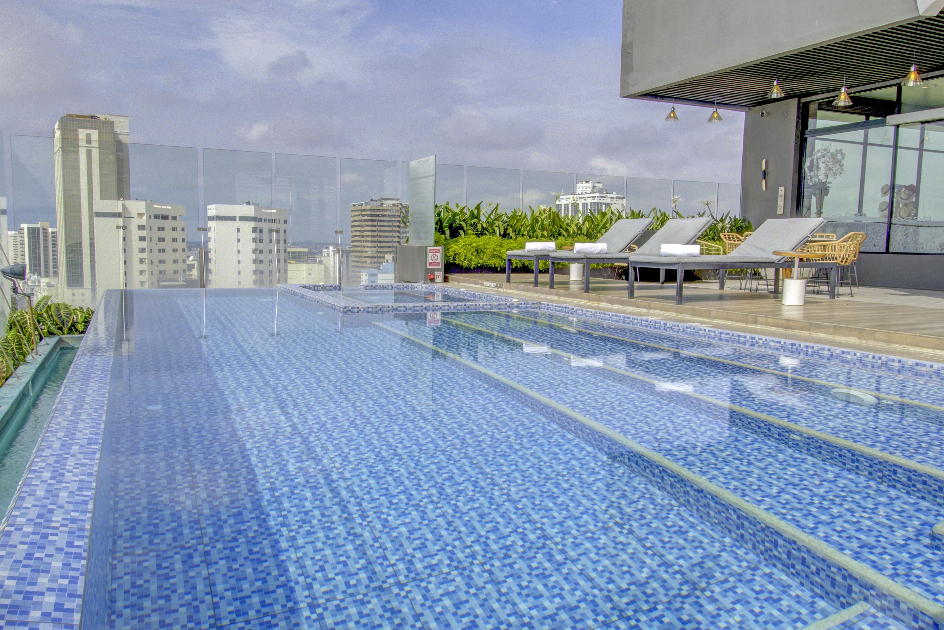 Piscina infinita en la azotea con sillones de descanso, rodeada de edificios de la ciudad.