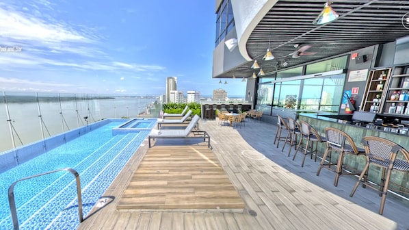 Área de piscina en la azotea con sillones de descanso, un bar y vistas al horizonte de la ciudad.