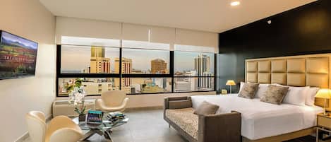 Una habitación de hotel moderna con una cama grande, un sofá, una mesa de centro de vidrio y una silla. Se observa una vista de la ciudad a través de amplias ventanas.