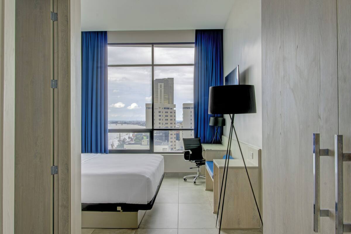 Ein Hotelzimmer mit Bett, Schreibtisch, Stuhl und einem großen Fenster mit Vorhängen.