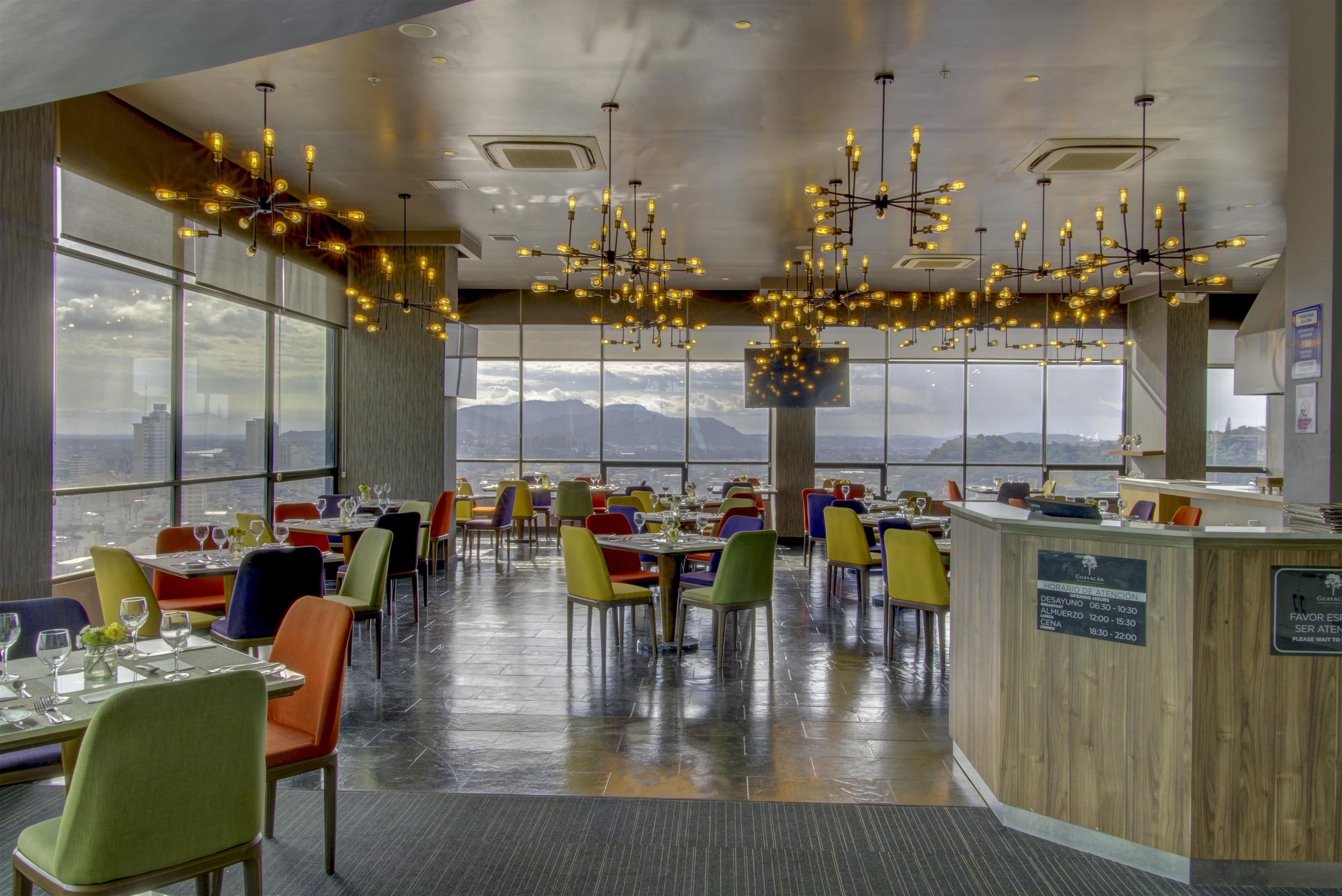 A restaurant with a high ceiling, multiple hanging light fixtures, and large windows offering a view of the cityscape.