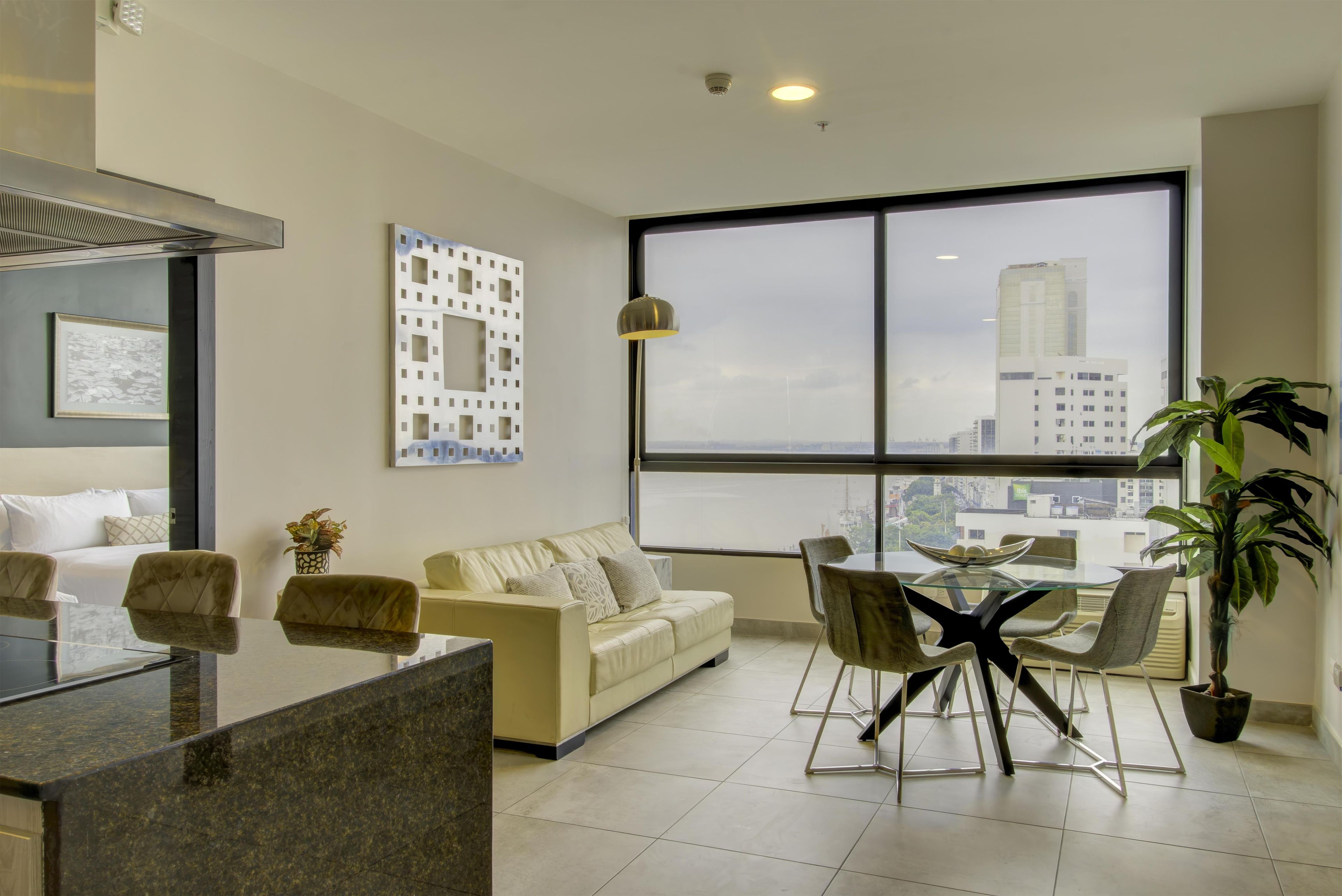 Una sala moderna con sofá, una mesa de comedor con sillas, un ventanal con vista a la ciudad y una zona de cocina con mostrador.