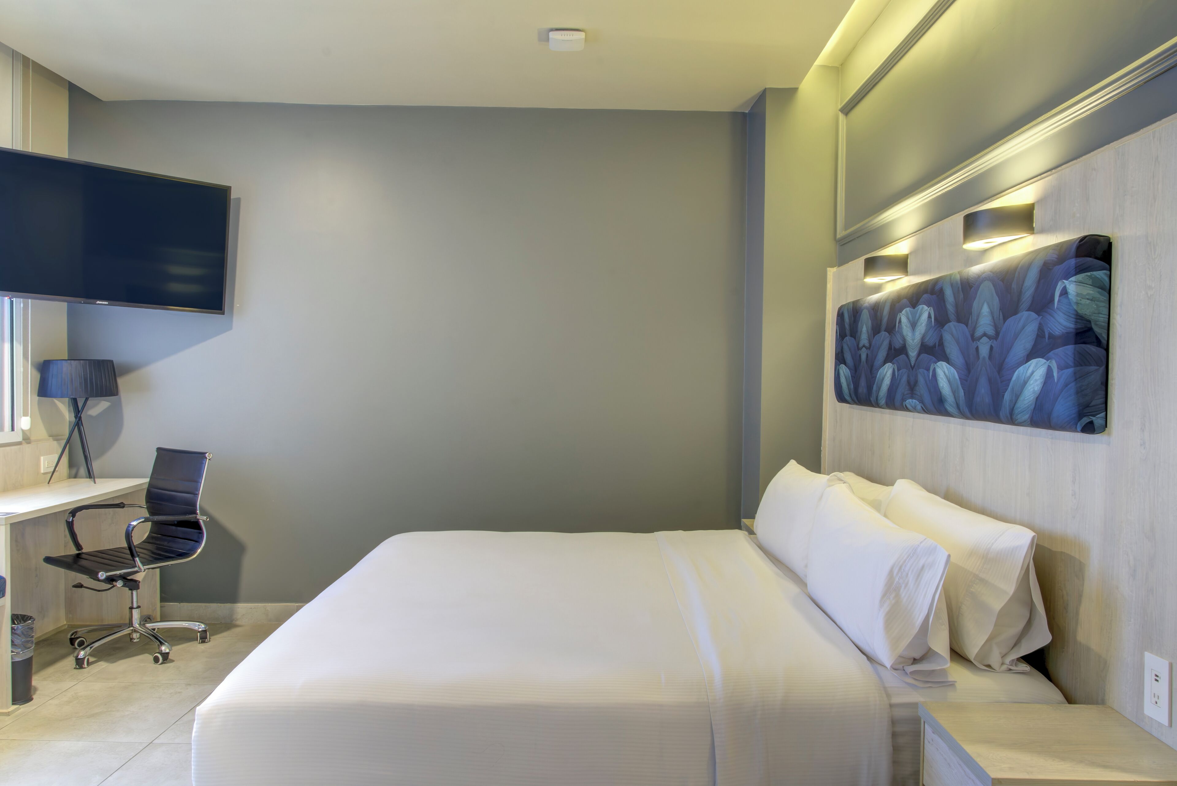 A hotel room with a bed, a desk with a chair, a flat-screen TV, and a painting on the wall.