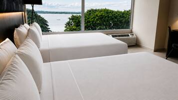 A modern hotel room with two beds, a large window with a view of a body of water and greenery, and an air conditioning unit.