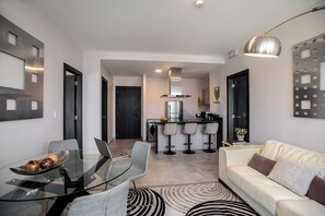 En moderne stue med et glasbord, grå stole, en cremefarvet sofa og et køkken i baggrunden.