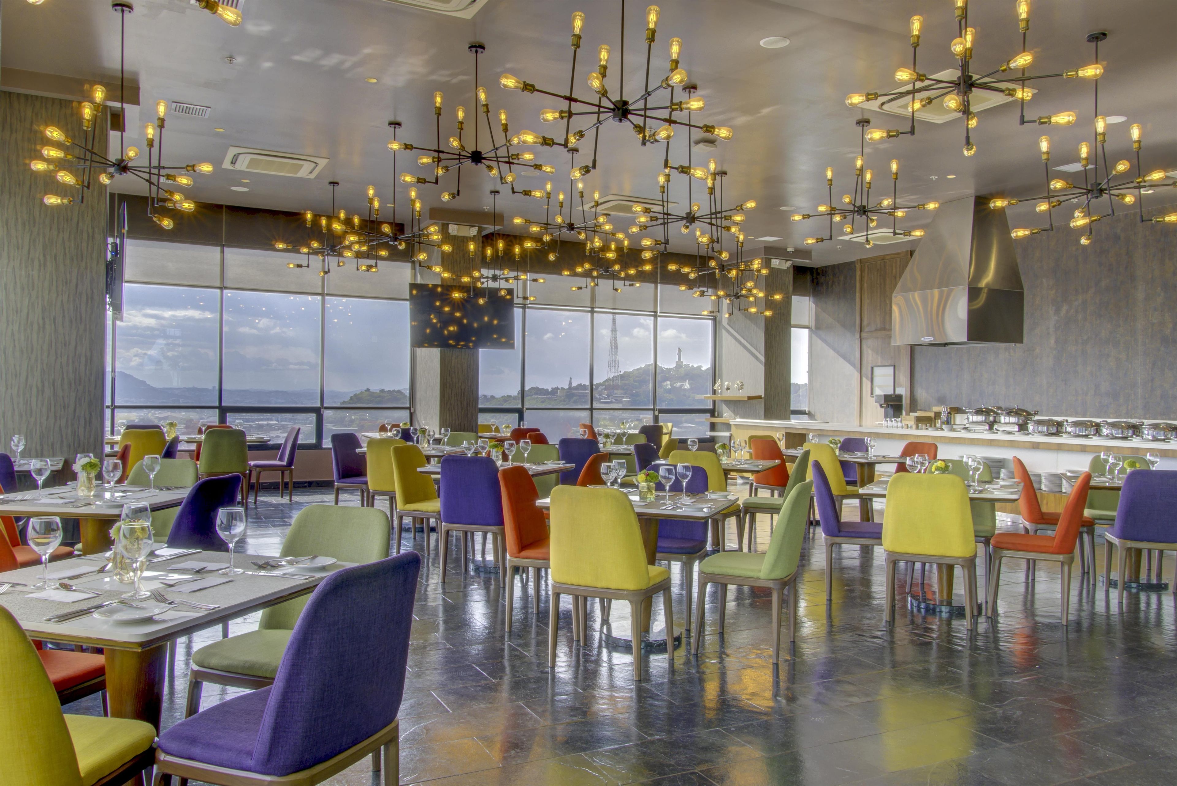 Un comedor amplio con varias mesas y sillas, ventanas grandes que ofrecen vista a la ciudad y un candelabro moderno con bombillas expuestas.