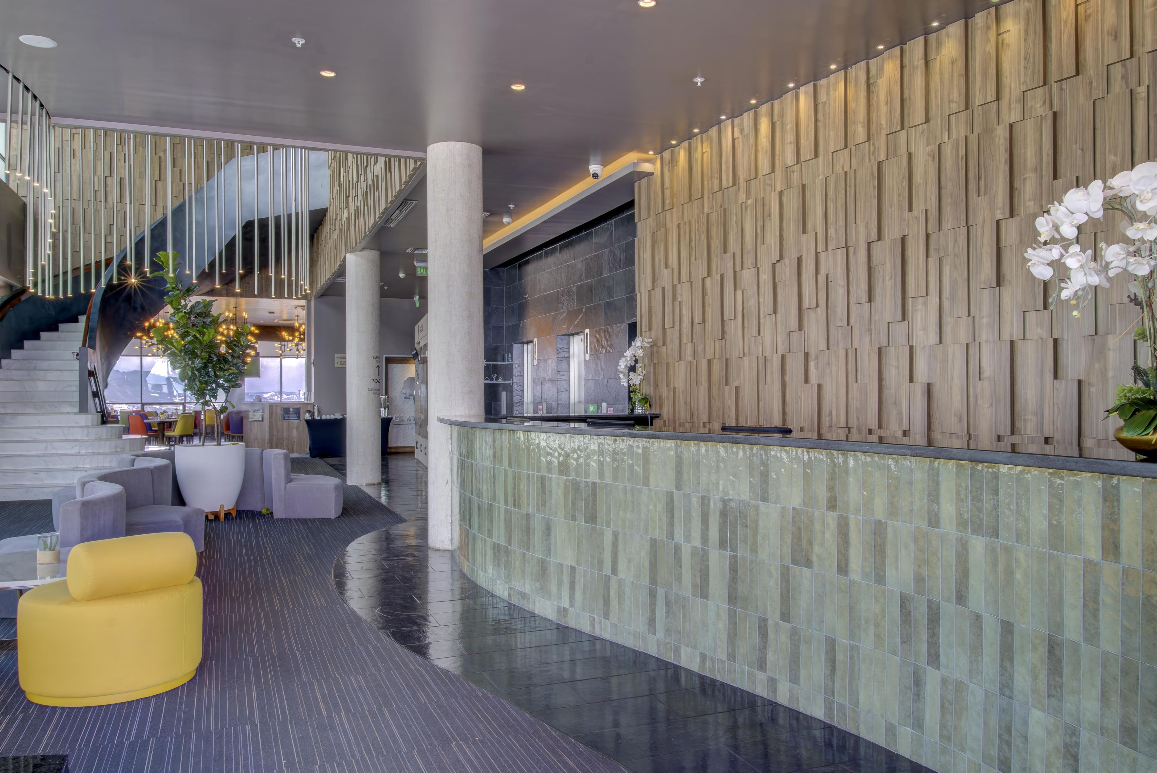 A modern lobby with a staircase, seating area, reception desk, and decorative plants.