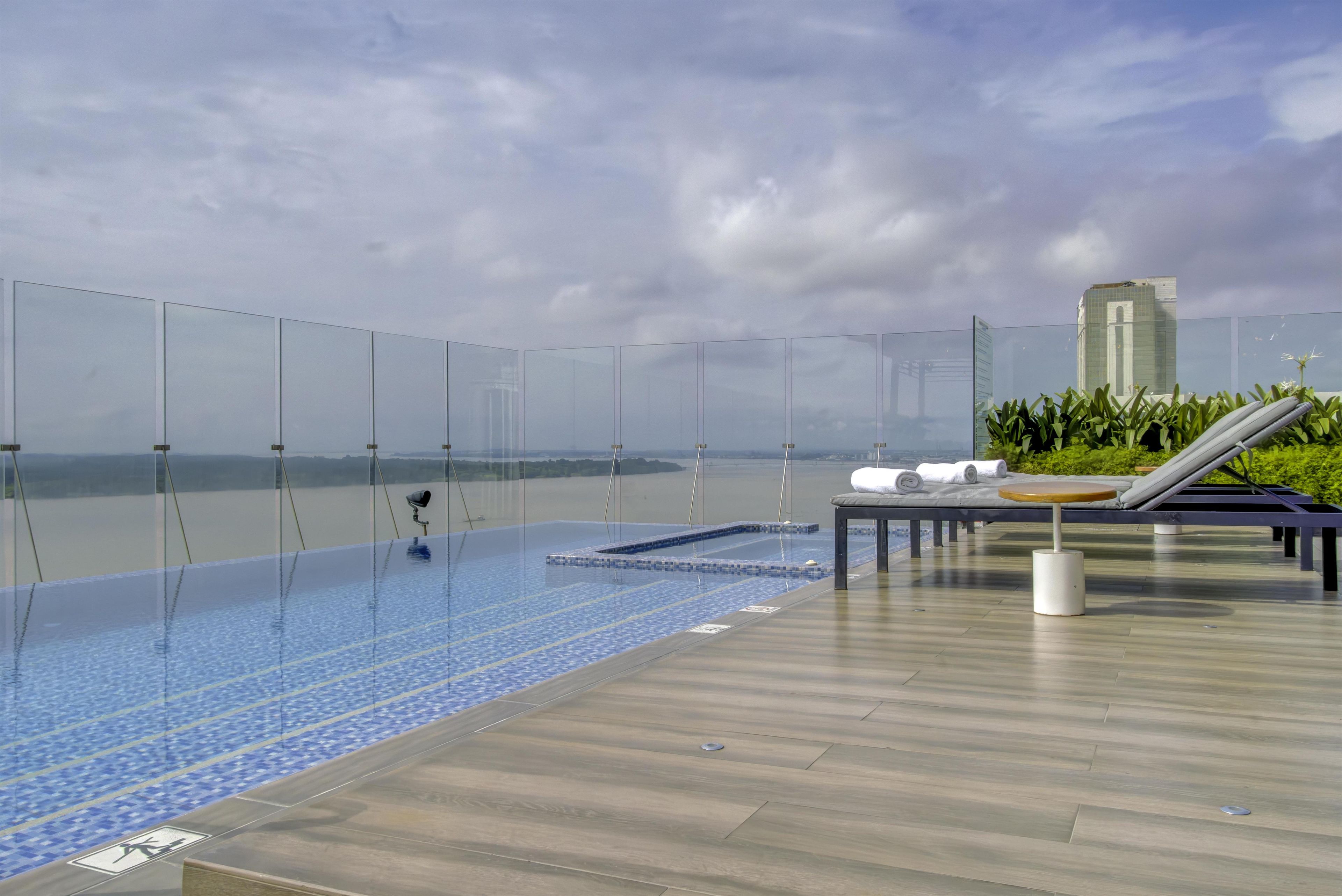 Una piscina infinita en la azotea con barandilla de cristal, un área de descanso con mesa y sillas, y vistas al horizonte de la ciudad.