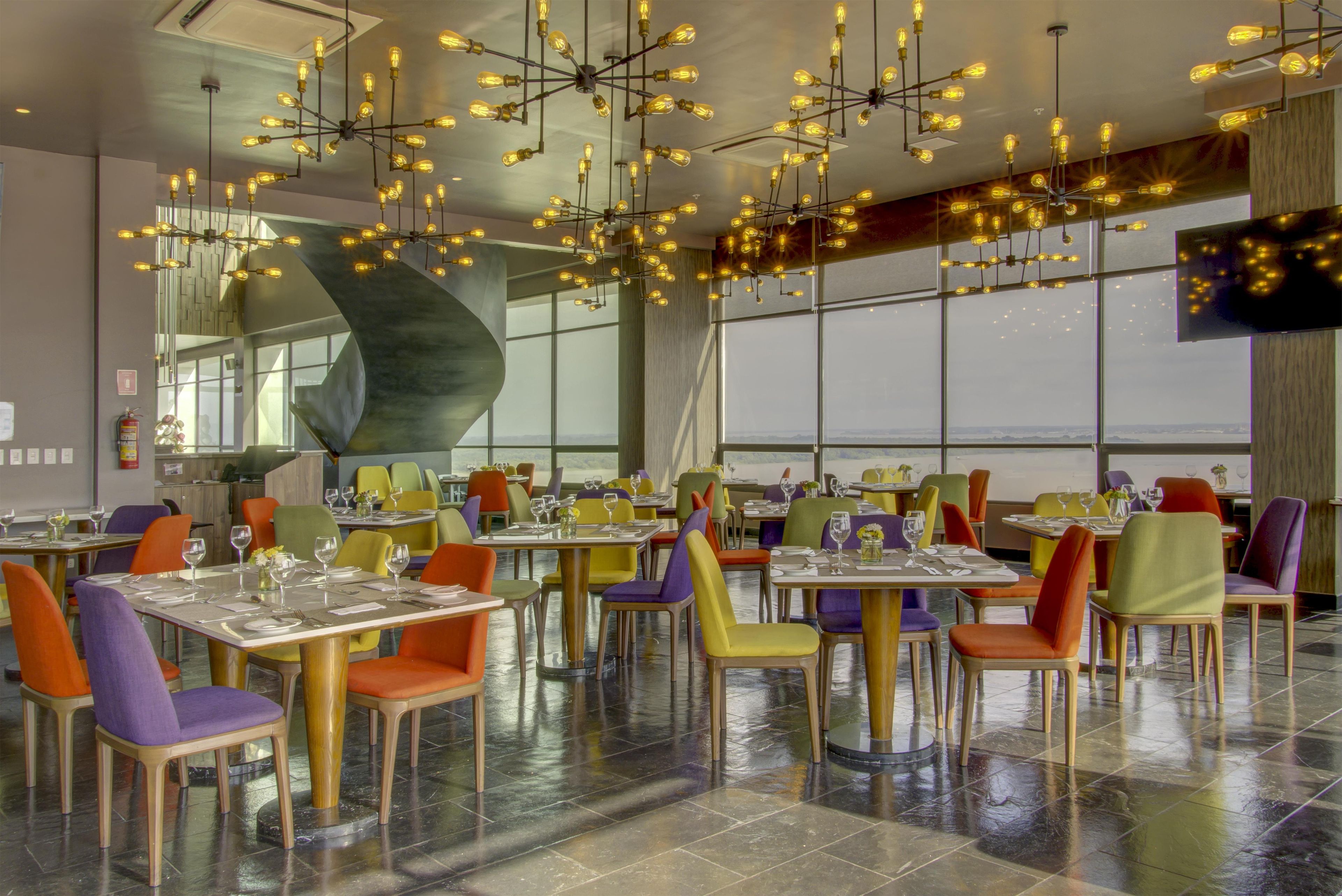 Ein Restaurant mit modernem Dekor, mit bunten Stühlen und Tischen, einer auffälligen Deckenleuchte und großen Fenstern mit Blick.