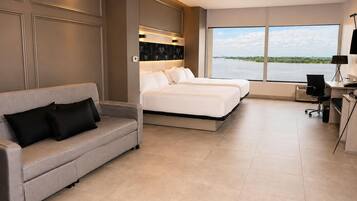 Habitación de hotel moderna con sofá, dos camas, un escritorio y un amplio ventanal con vista al agua.