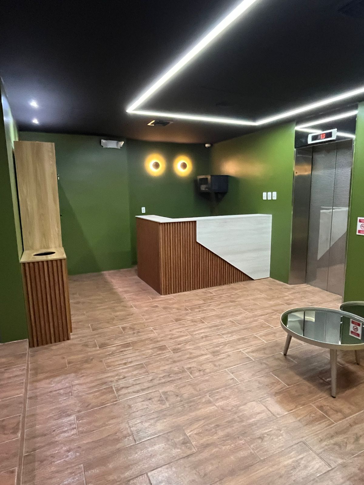 Un espace d’accueil avec un comptoir en bois, une table en verre et un écran numérique indiquant l’étage de l’ascenseur.