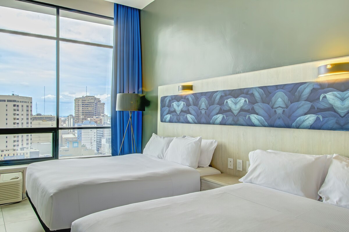 Hotelkamer met twee bedden, een groot raam met uitzicht op de stad en een wand met een kunstwerk met een bladmotief.