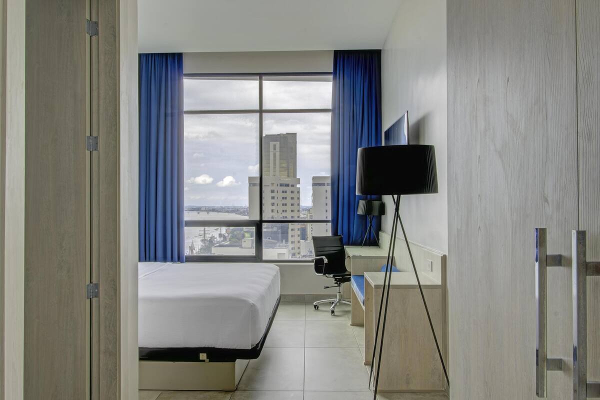 Ein Hotelzimmer mit Bett, Schreibtisch, Stuhl und einem großen Fenster mit Vorhängen.