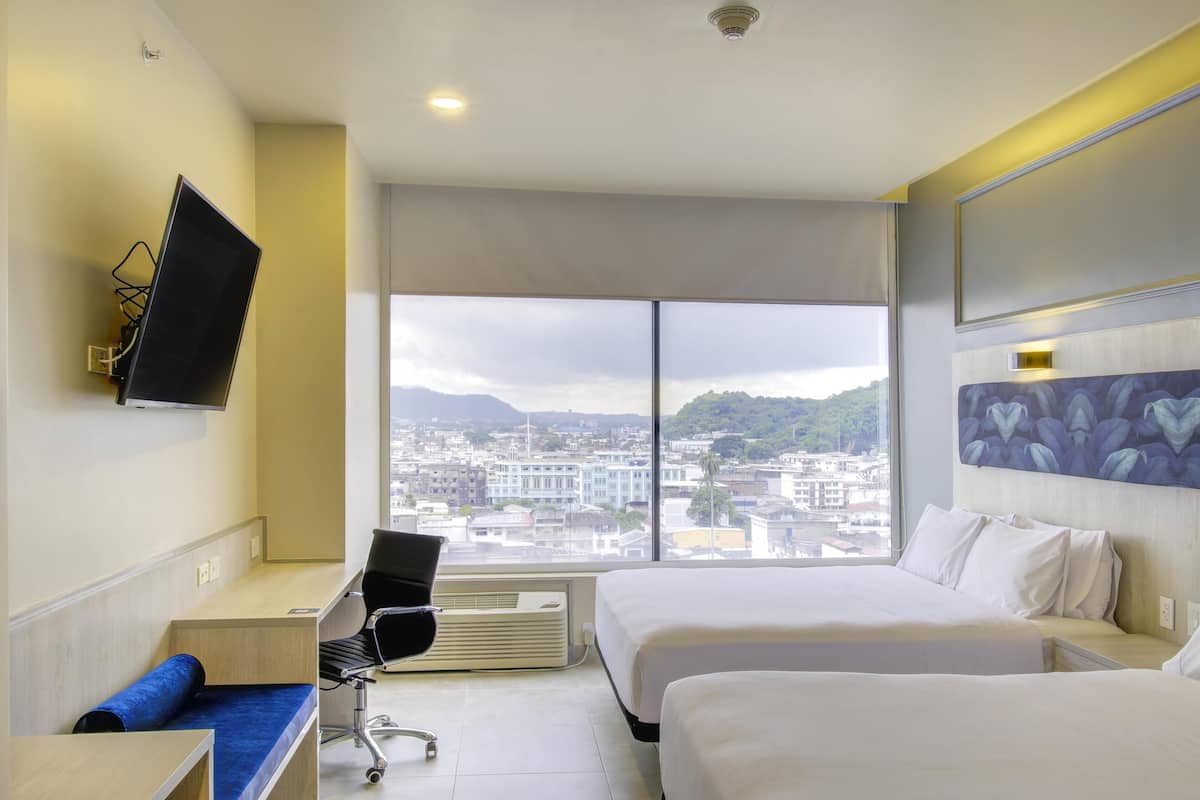 Ein Hotelzimmer mit zwei Betten, einem Schreibtisch, einem Stuhl, einem wandmontierten Flachbildfernseher und einem großen Fenster mit Blick auf die Stadt.