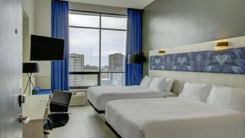 A hotel room with two beds, a desk with a chair, a TV, and a large window with a city view.