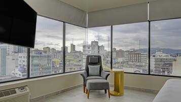 A modern room with a large window, a grey armchair with a cushion, a small round table, and a bed.
