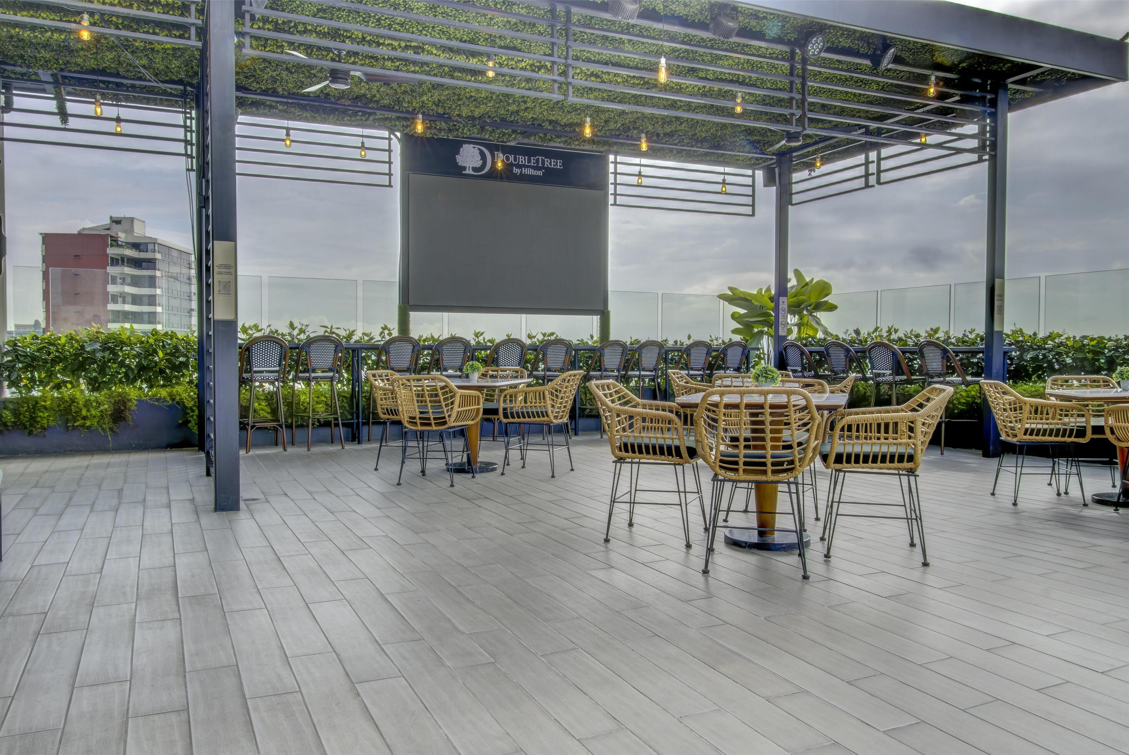 An outdoor seating area with wicker chairs and tables, a large screen, and a pergola with string lights.