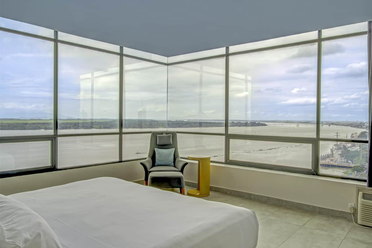 Ein Hotelzimmer mit einem großen Bett, einem Sessel und einem kleinen Tisch. Große Fenster bieten Blick auf eine ferne Landschaft.