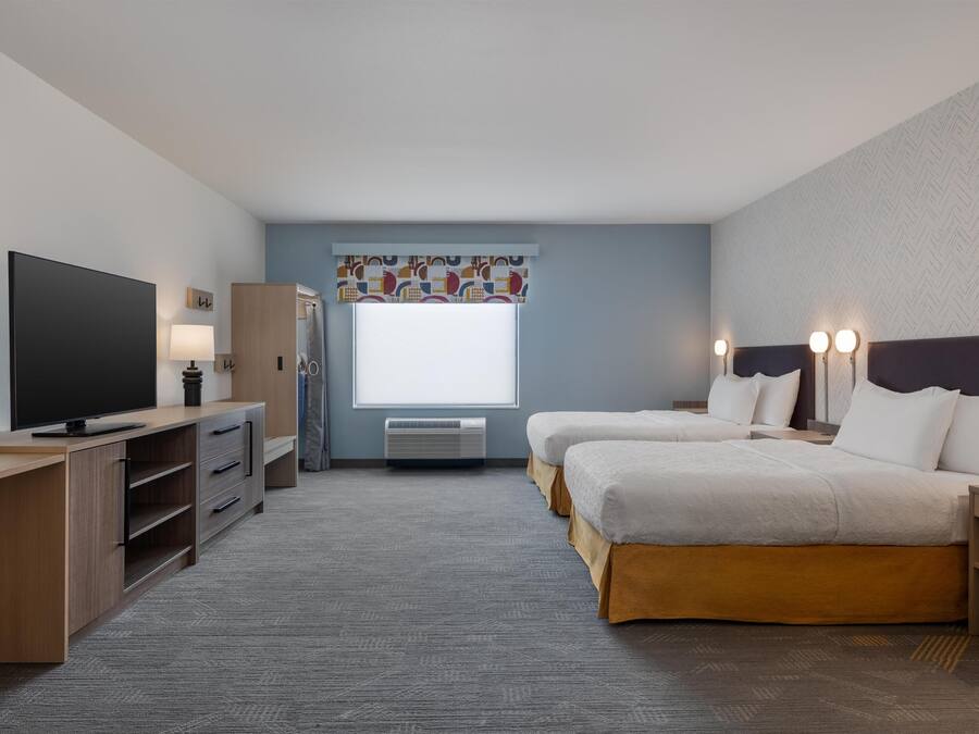 A hotel room with a flat-screen TV, a desk with drawers, a window with blinds, two beds with headboards, and a wall-mounted air conditioning unit.