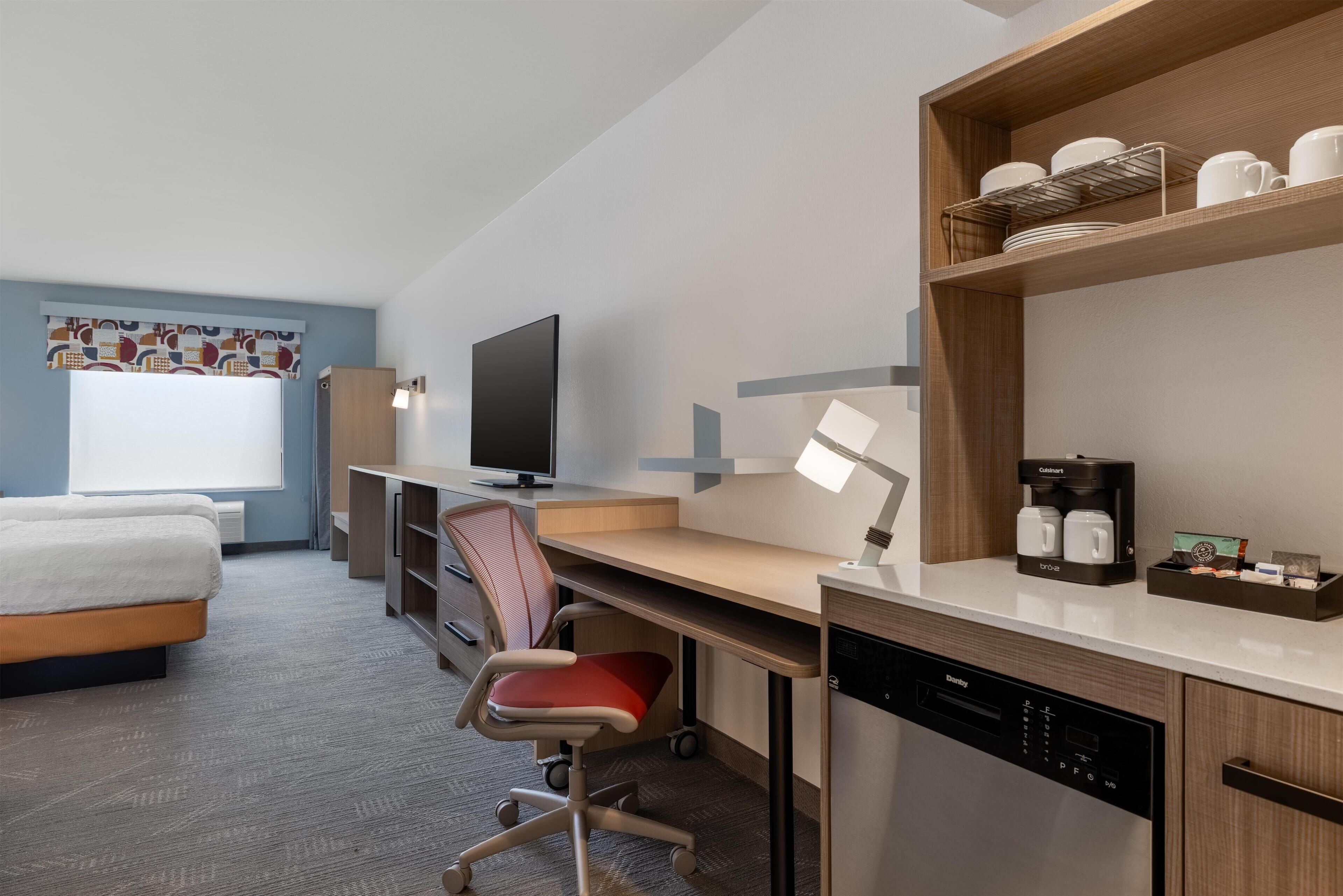 A modern hotel room with a bed, desk, chair, television, coffee maker, and a dishwasher.