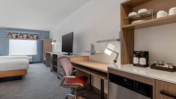 A modern hotel room with a bed, desk, chair, television, coffee maker, and a dishwasher.