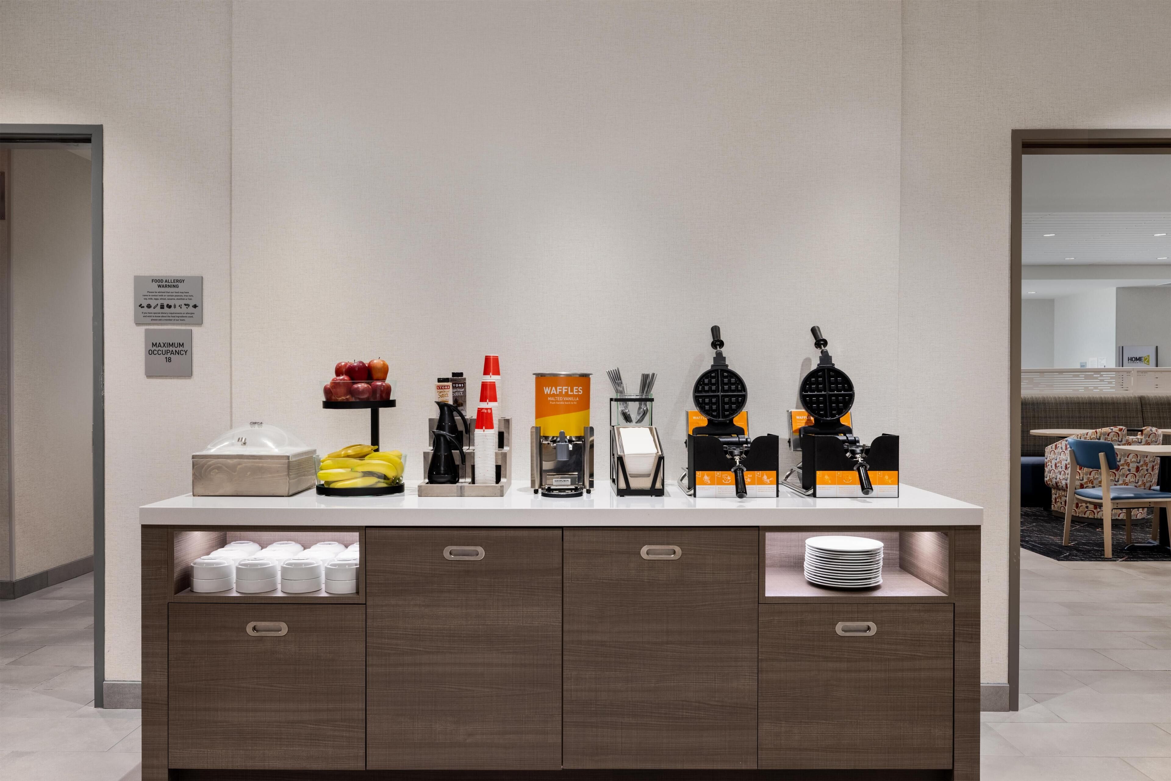 A breakfast bar with various food items and appliances, including a waffle maker, coffee maker, and a sign indicating a maximum occupancy limit.
