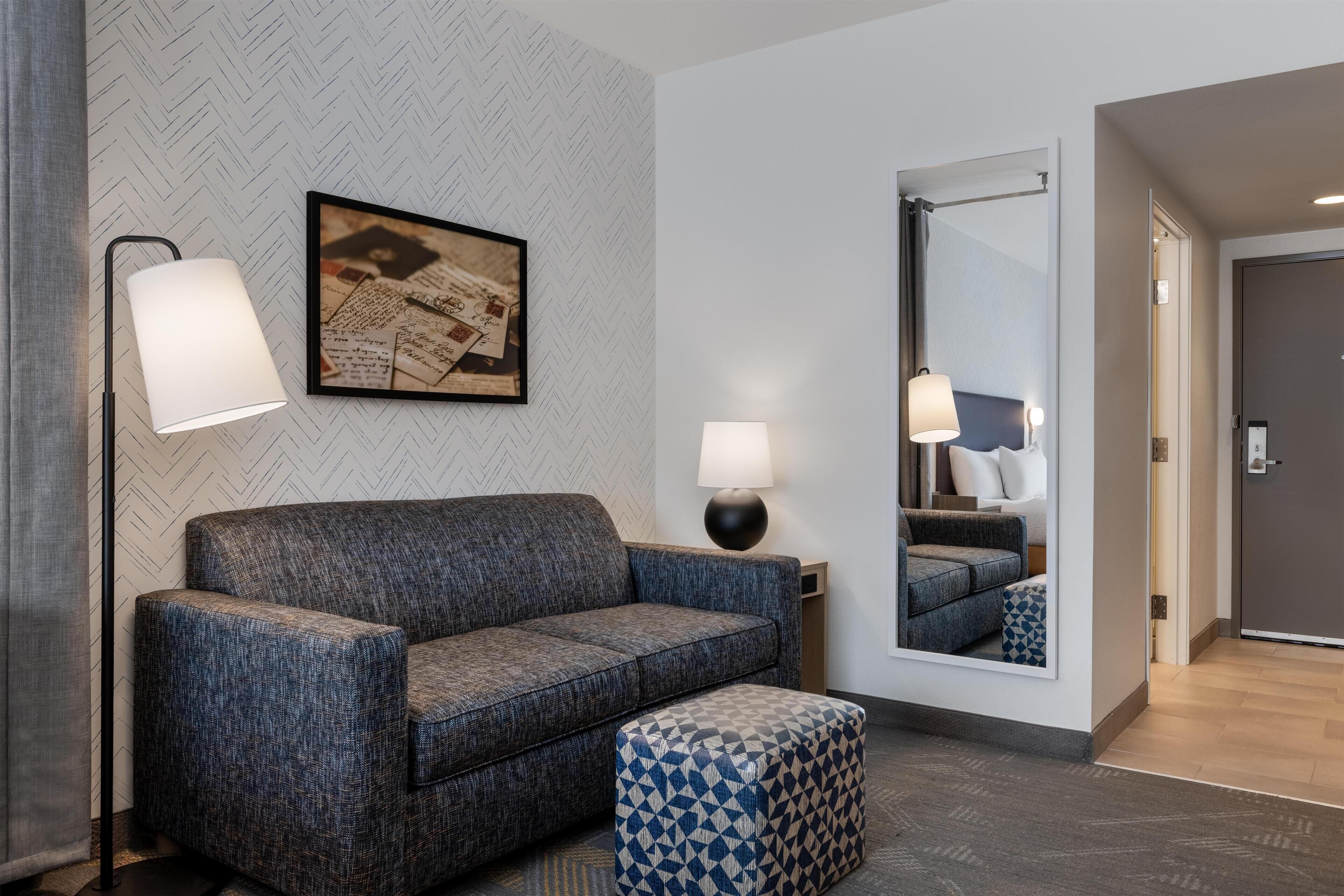 A modern hotel room with a grey herringbone-patterned wall, a grey sofa, a patterned ottoman, two bedside lamps, and a large mirror reflecting the room.