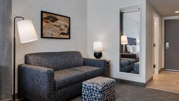 A modern hotel room with a grey herringbone-patterned wall, a grey sofa, a patterned ottoman, two bedside lamps, and a large mirror reflecting the room.