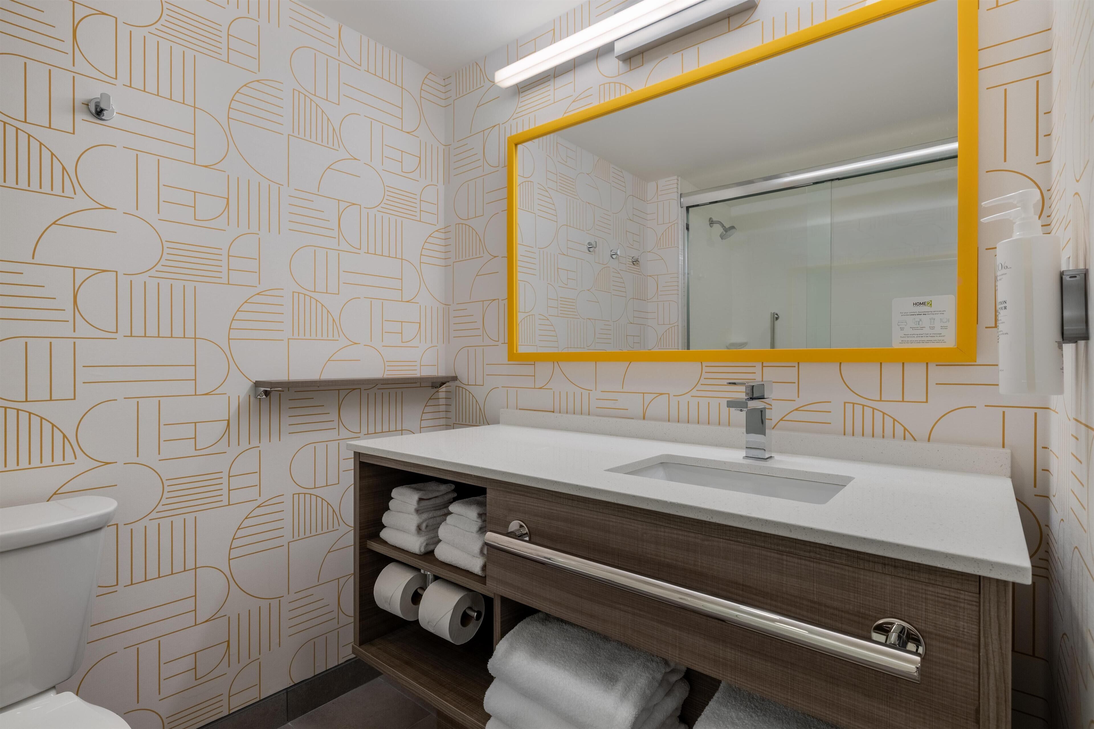 Une salle de bain moderne avec un mur orné d’un motif géométrique, un lavabo blanc avec un meuble-vasque en bois et un miroir avec un cadre jaune.