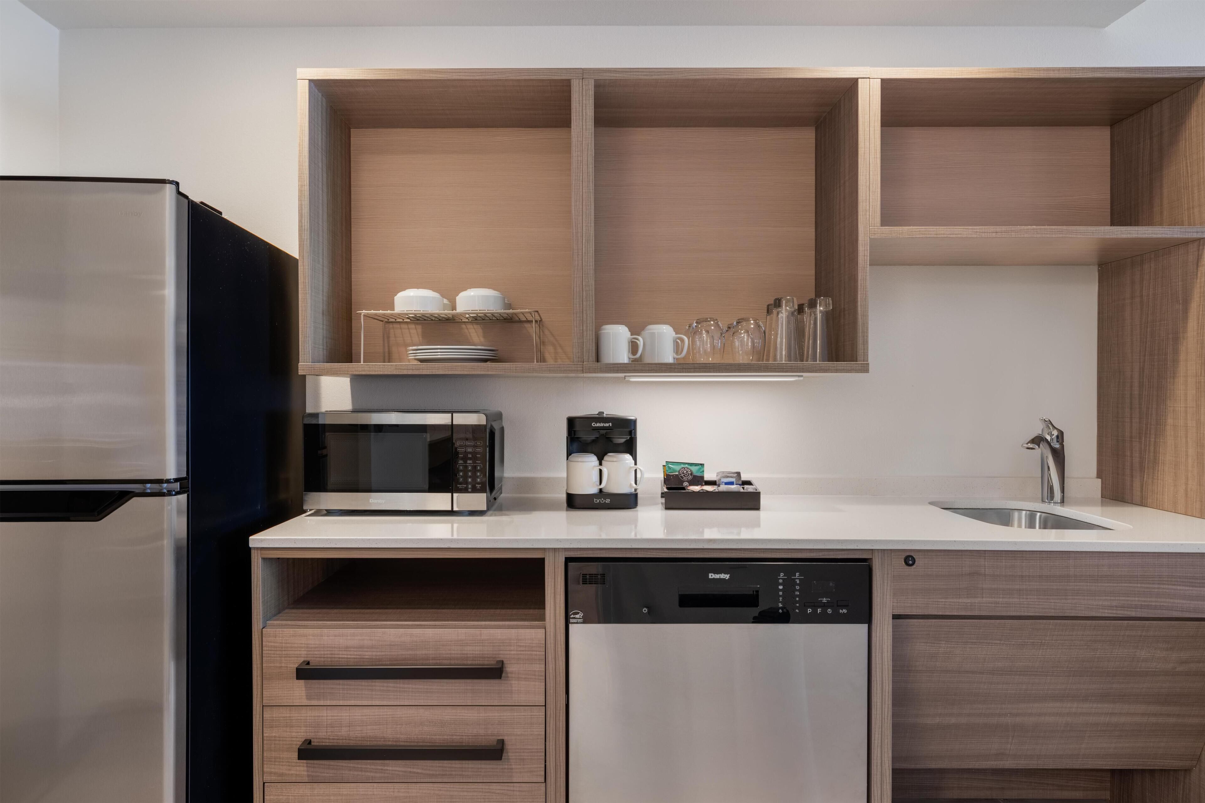 Una cocina moderna con un refrigerador de acero inoxidable, un microondas integrado y un lavavajillas. Hay armarios de madera y una encimera con una cafetera y tazas.