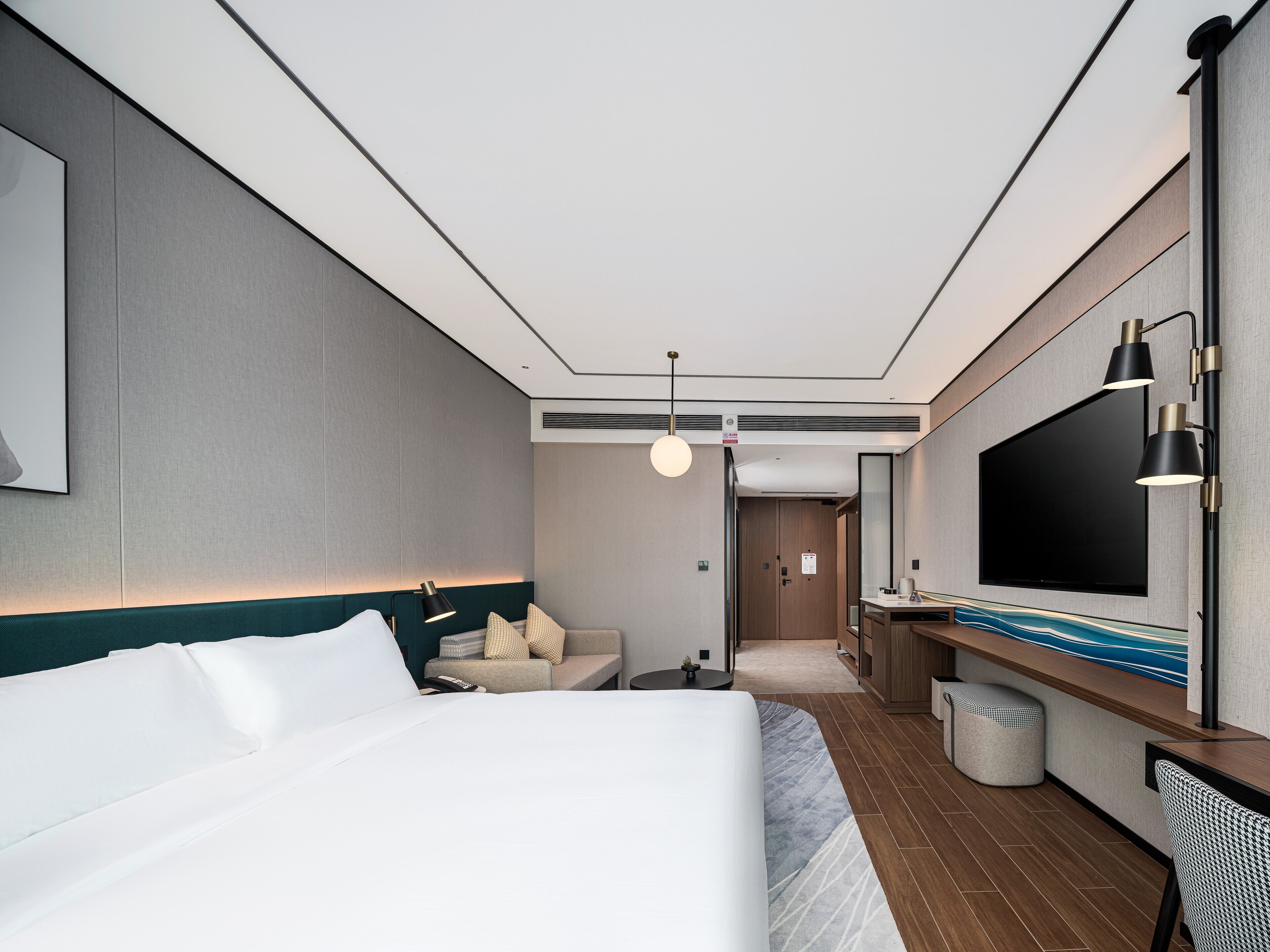 A modern hotel room with a large bed, a desk with a flat-screen TV, a seating area, and a hallway leading to another room.