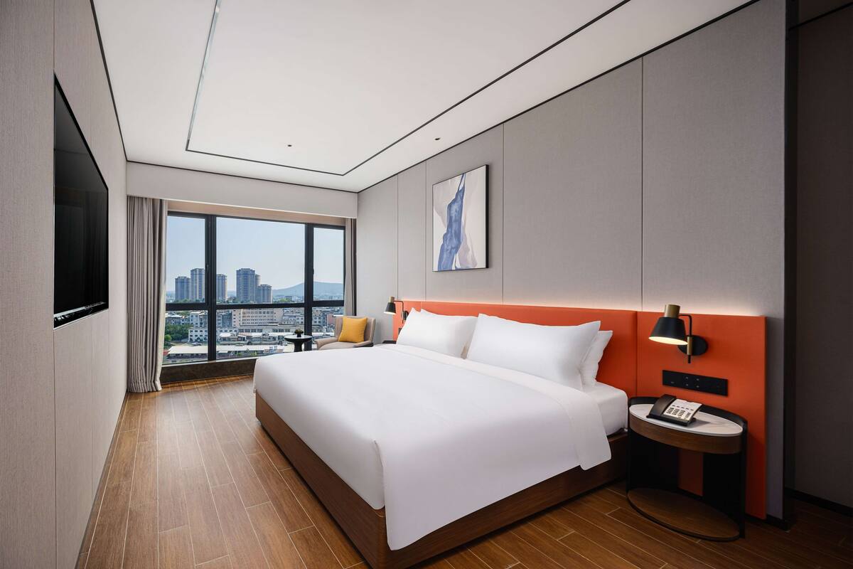 A modern hotel room with a large bed, a city view, a flat-screen TV, and a bedside table with a lamp.