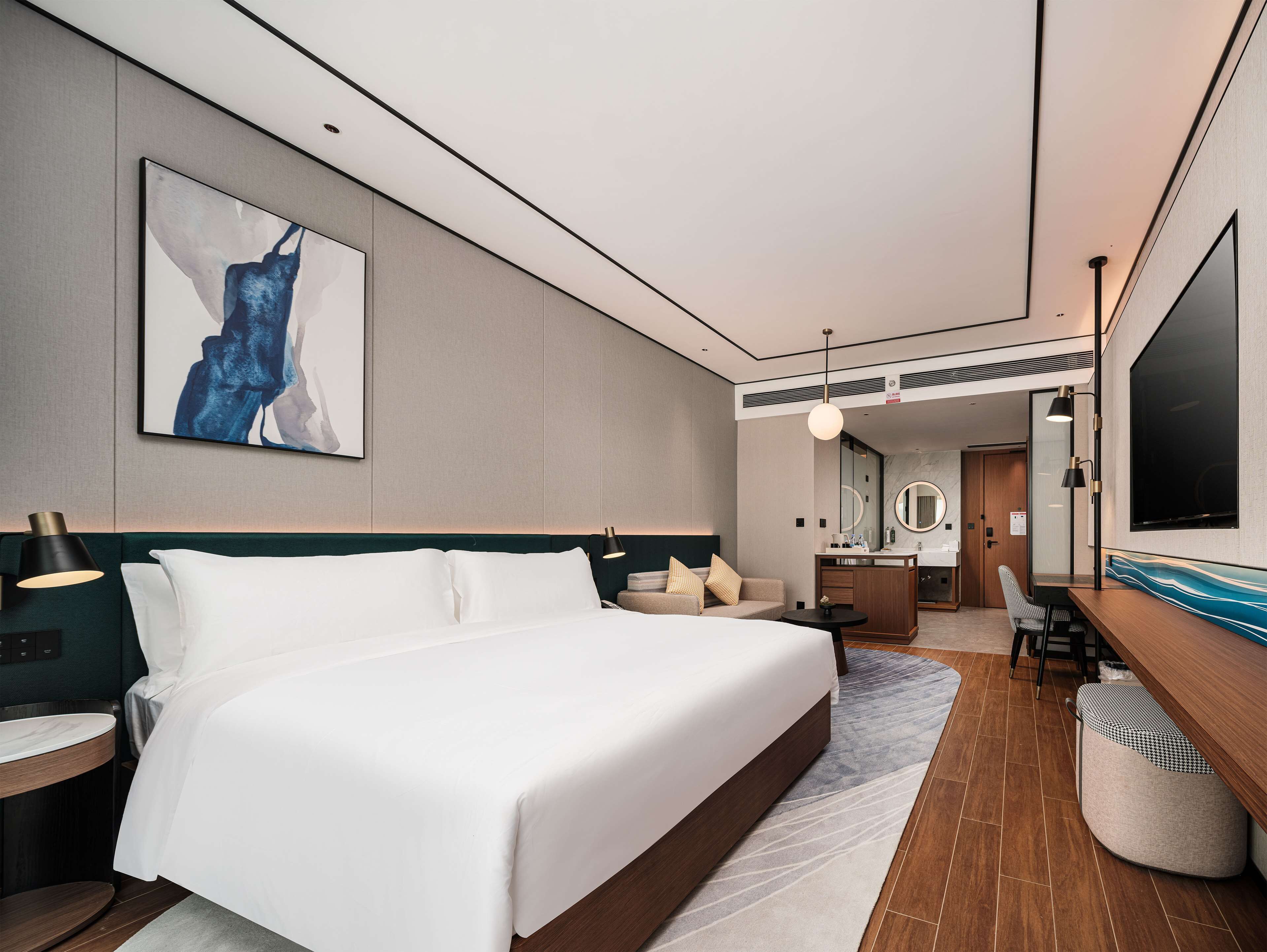 A modern hotel room with a large bed, a desk with a lamp, a vanity with a mirror, and a TV.