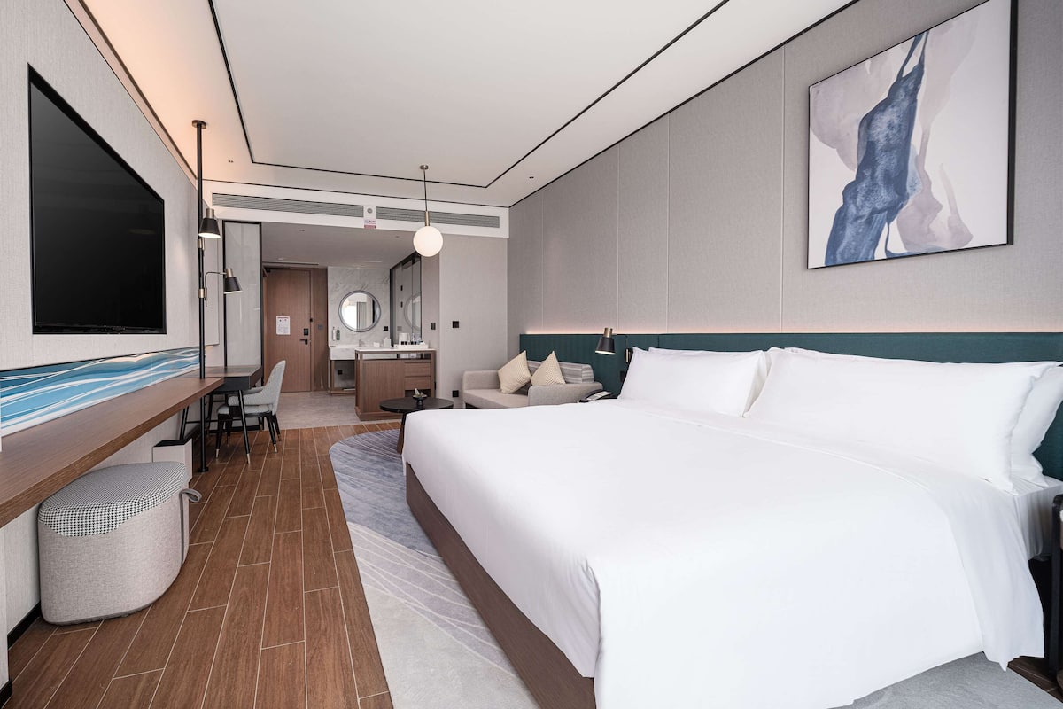 A modern hotel room with a large bed, a desk with a chair, a television, and a bathroom with a mirror.