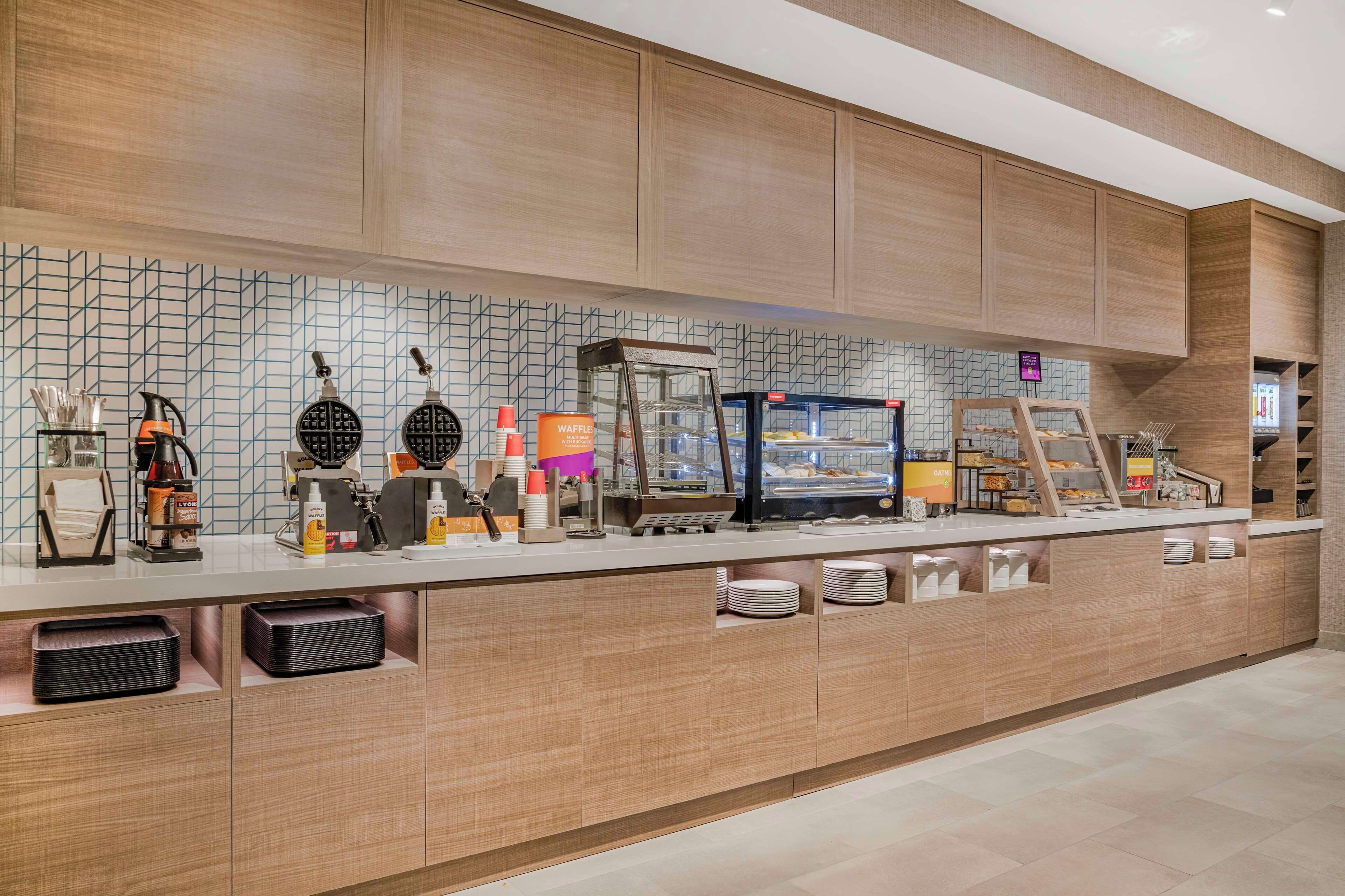 A modern breakfast bar with waffle makers, a coffee machine, and a refrigerated display.