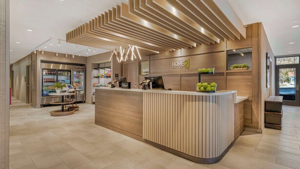 A modern hotel reception area with a wooden reception desk, a digital kiosk, and a refrigerator stocked with beverages. - Home2 Suites By Hilton Boone (Boone)