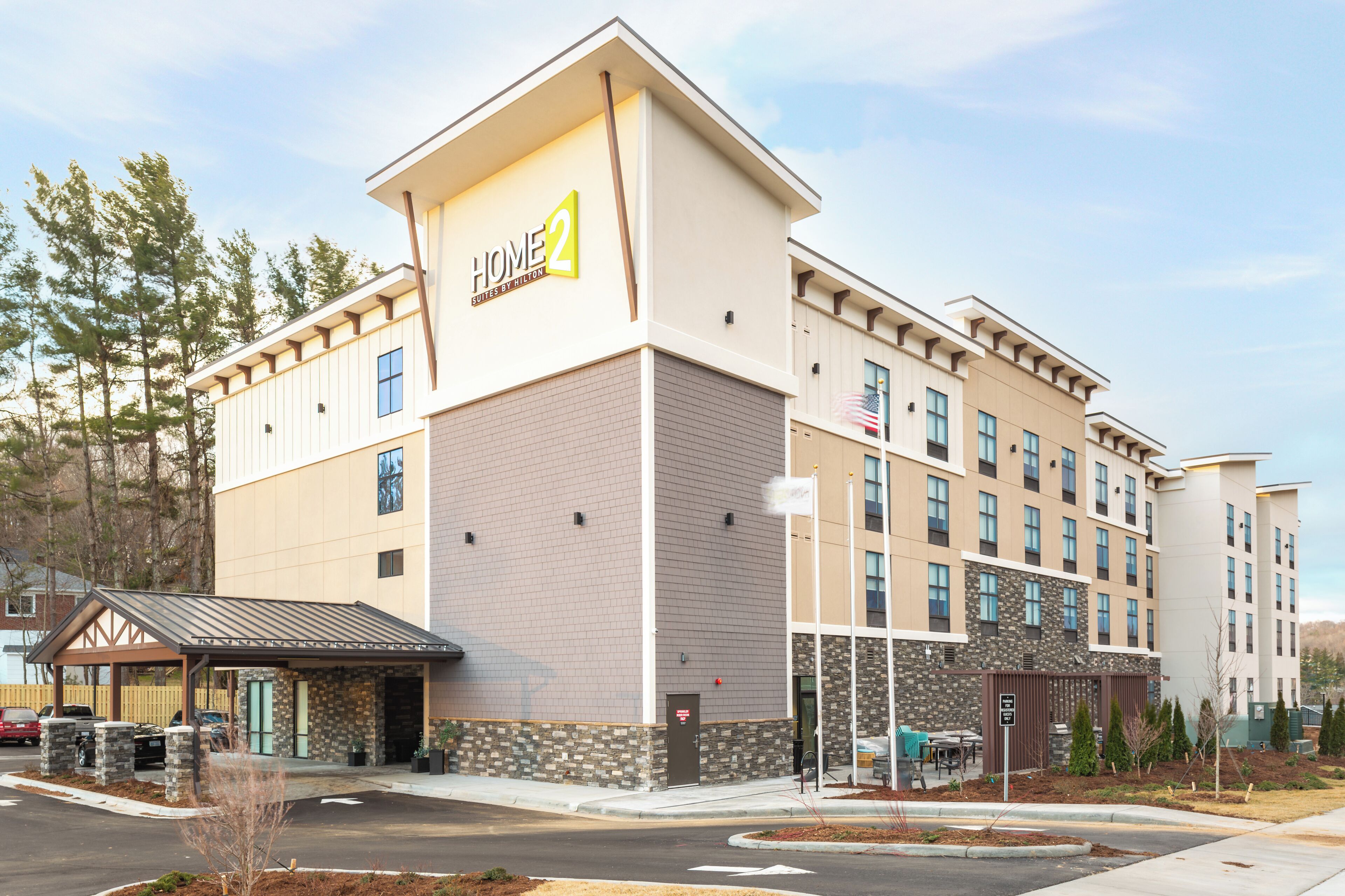 Un hotel de varios pisos con un cartel llamativo de 'Home 2 Suites by Hilton', una entrada cubierta y un estacionamiento con coches.