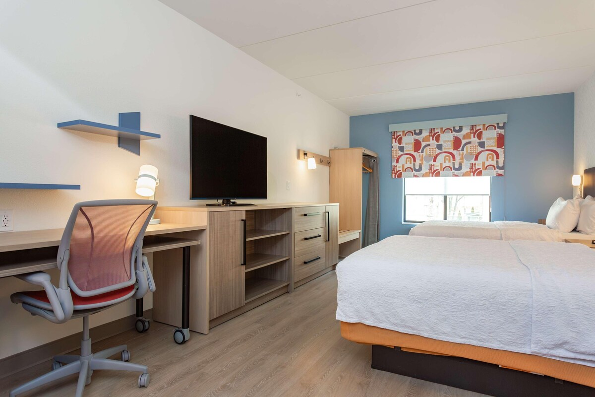 A hotel room with a bed, desk, chair, television, wardrobe, and a window with curtains.