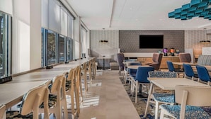 A modern dining area with long tables and chairs, a kitchen counter with a flat-screen TV, and a patterned wall. - Home2 Suites By Hilton Boone (Boone)