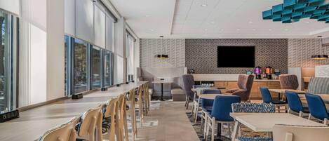 A modern dining area with long tables and chairs, a kitchen counter with a flat-screen TV, and a patterned wall.