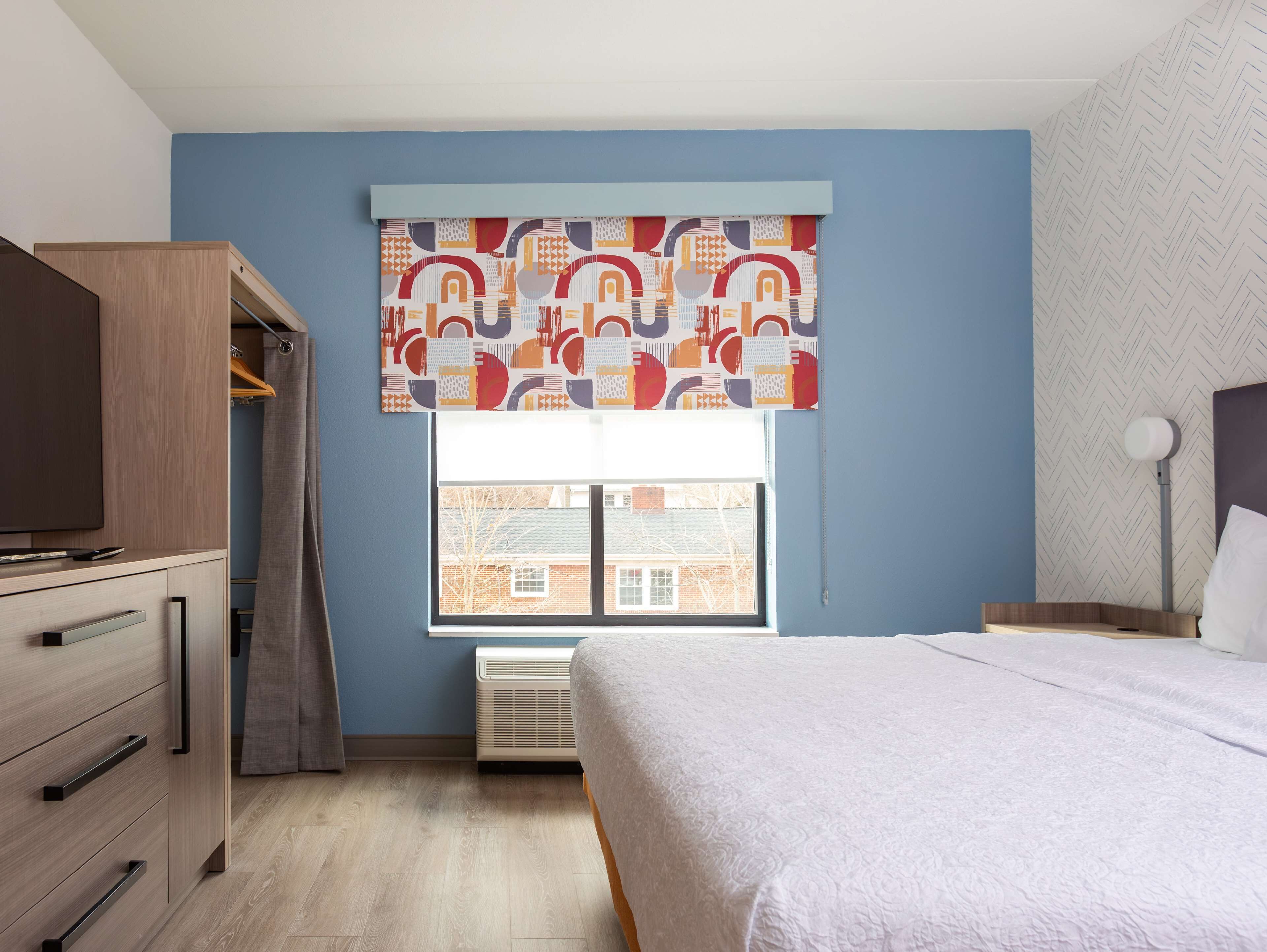 A bedroom with a bed, a wardrobe, a desk, a window with a view of buildings, and a patterned curtain.