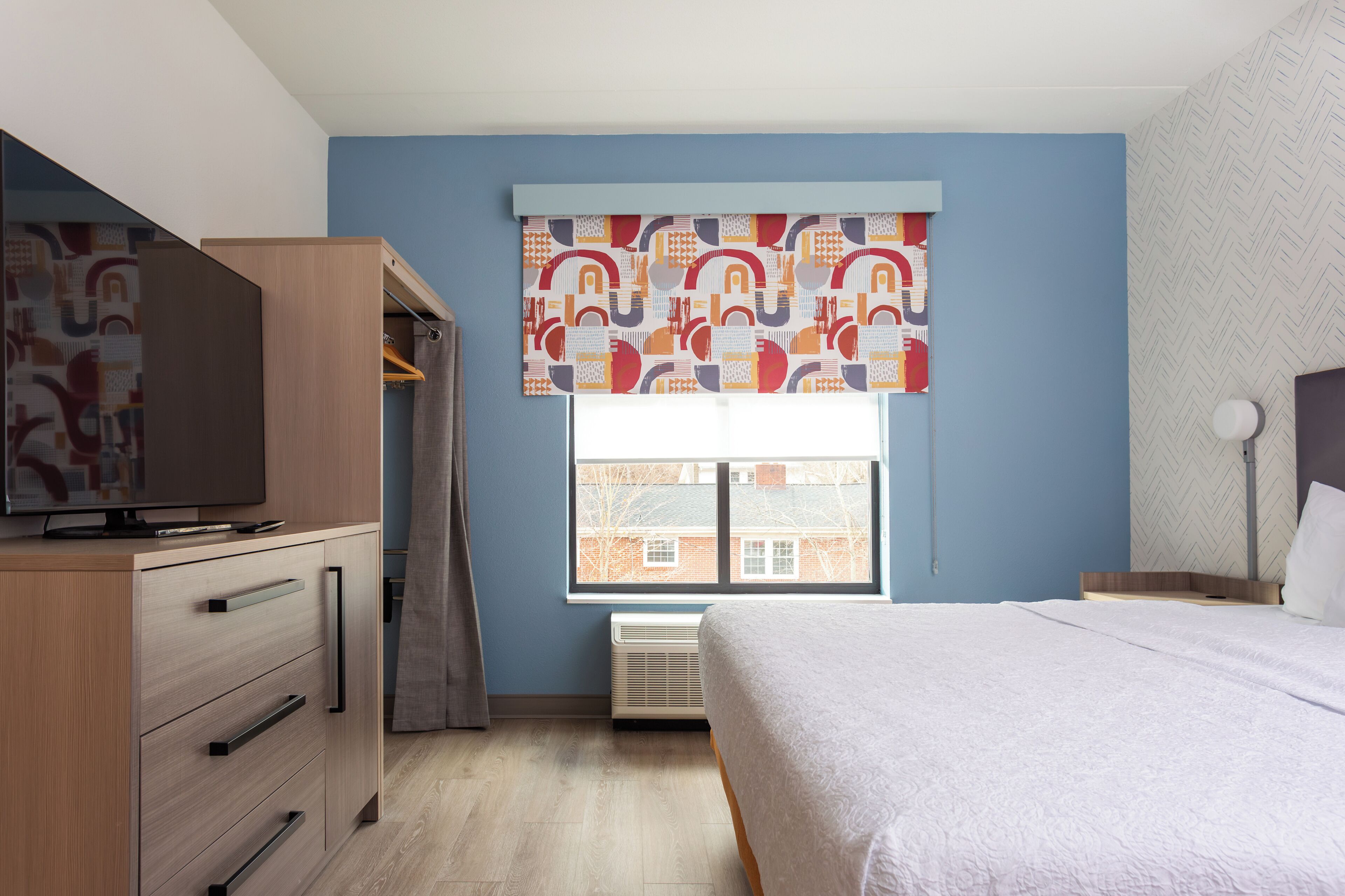 A bedroom with a bed, a wardrobe, a television, and a window with patterned curtains.