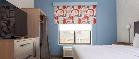 A bedroom with a bed, a wardrobe, a television, and a window with patterned curtains.