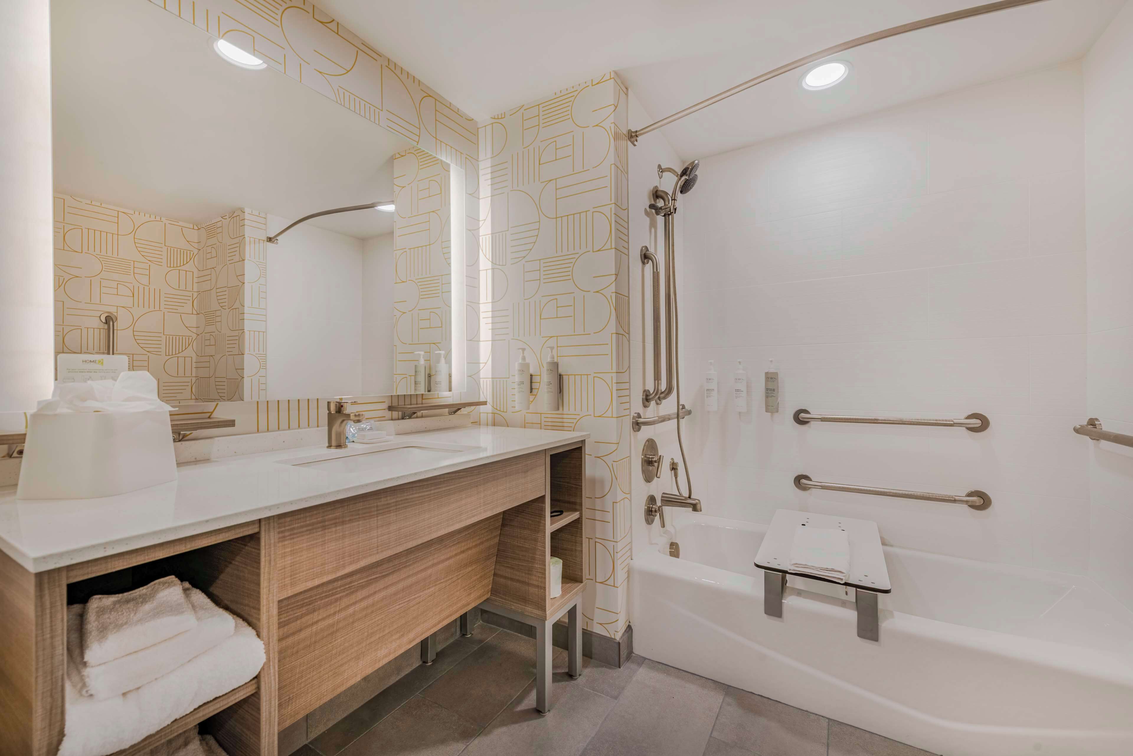 A modern bathroom with a large mirror, two sinks, a shower with grab bars, and a towel rack.